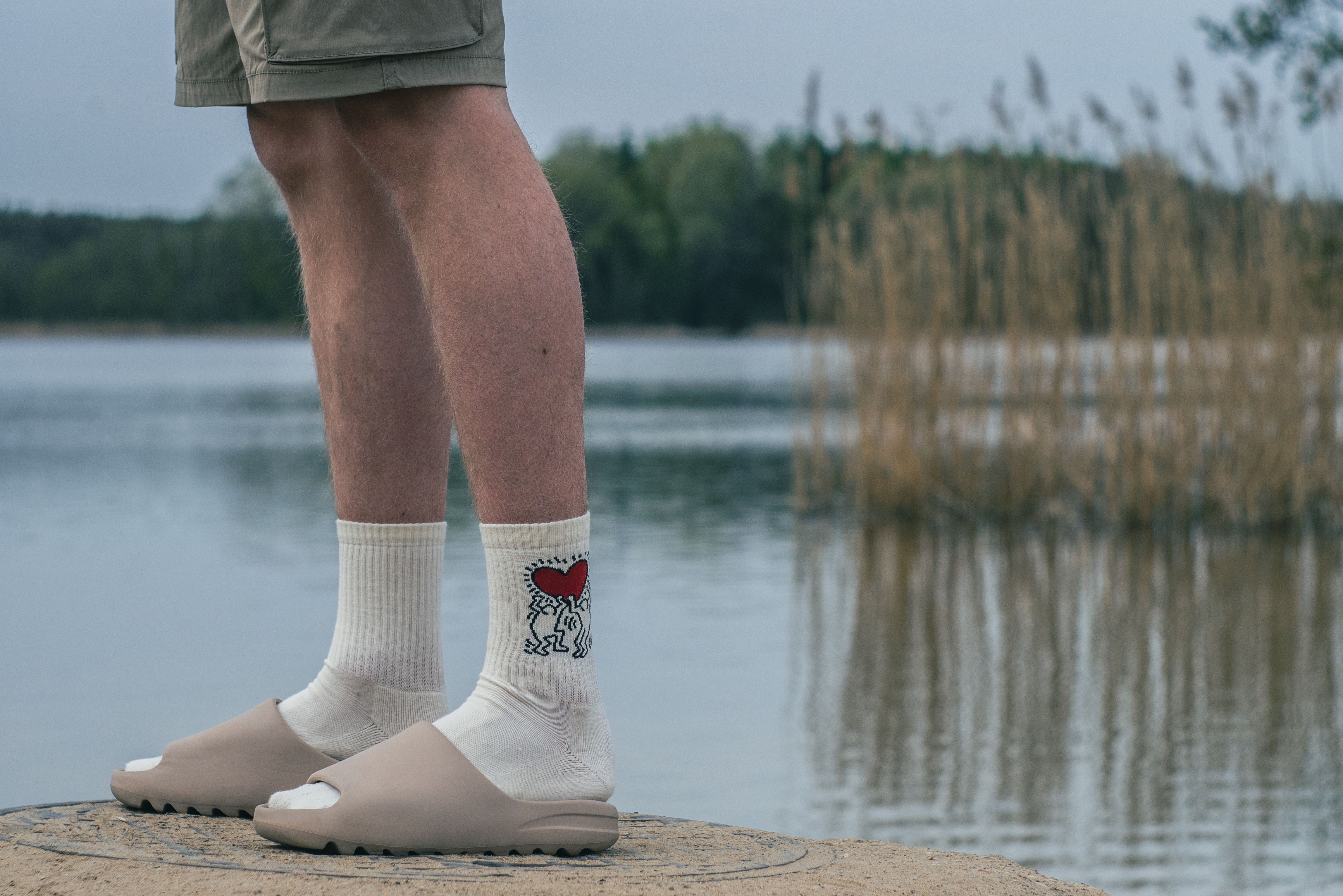 A person stands by a calm lake wearing casual shorts, logo socks, and slides, emphasizing leisure and nature.
