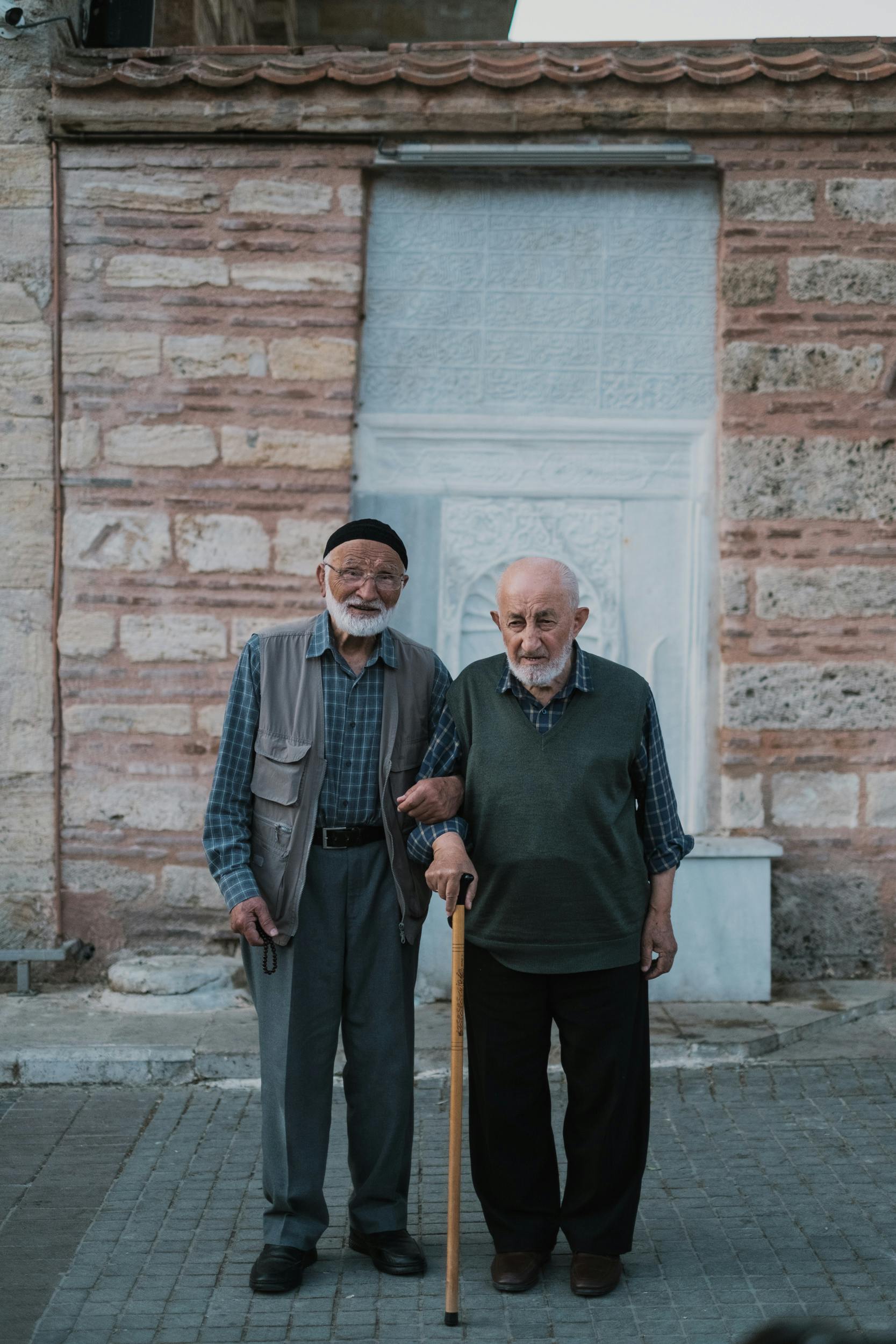 Two elderly men stand together on a historic street in Bursa, Türkiye, showcasing friendship.
