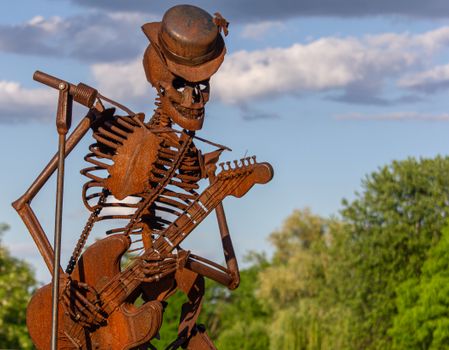 A rusty skeleton sculpture in the shape of a musician playing guitar outdoors under a blue sky.