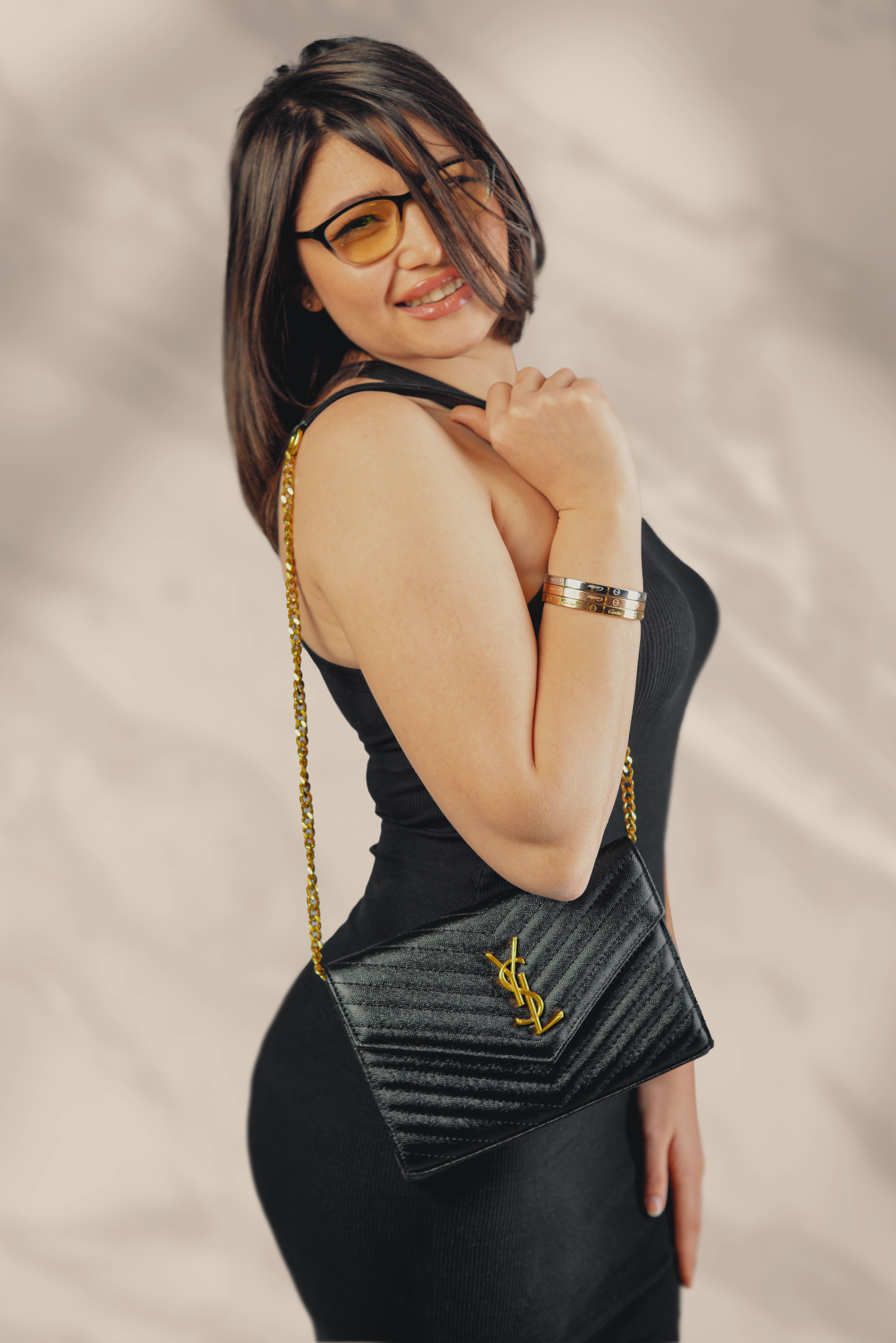 Stylish woman smiling in a black dress, showcasing a luxury YSL sling bag and sunglasses.
