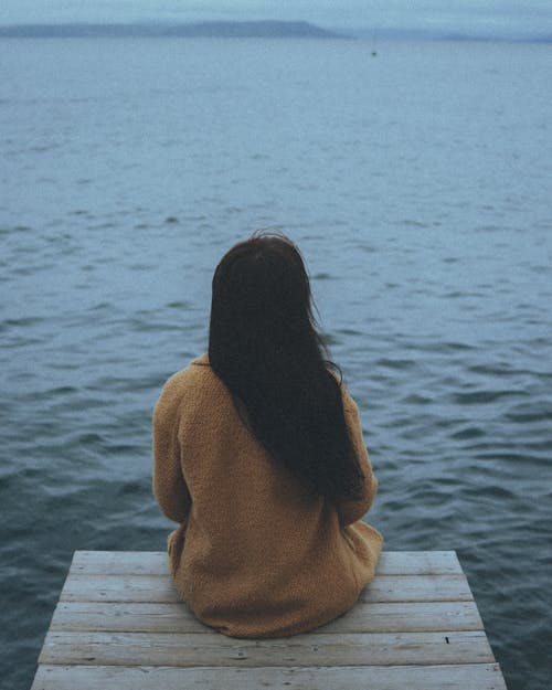 Free Woman in Brown Long Sleeve Shirt Sitting on White Wooden Bench Near Body of Water during Stock Photo