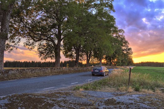 A car travels down a rural road lined with trees during a vibrant sunset.