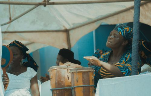 Free Elderly African woman drumming at a vibrant cultural festival under a canopy. Stock Photo