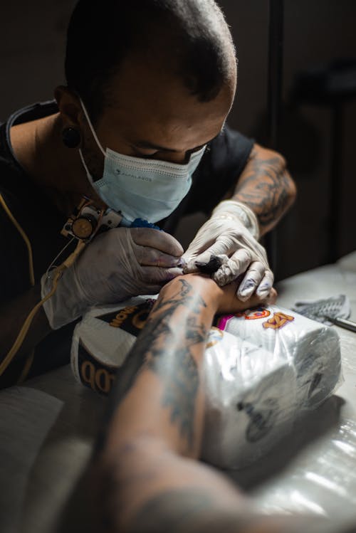 A Man in Black Shirt Wearing Gloves and Face Mask while Doing Tattoo
