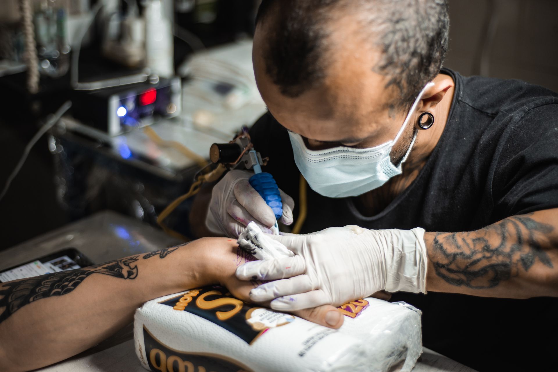 A masked tattoo artist wearing gloves uses a tattoo machine while working on a client’s tattooed forearm.
