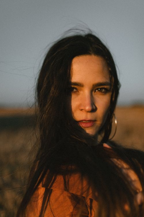 Free A captivating sunlit portrait of a woman with long brunette hair in a field, exuding confidence and elegance. Stock Photo