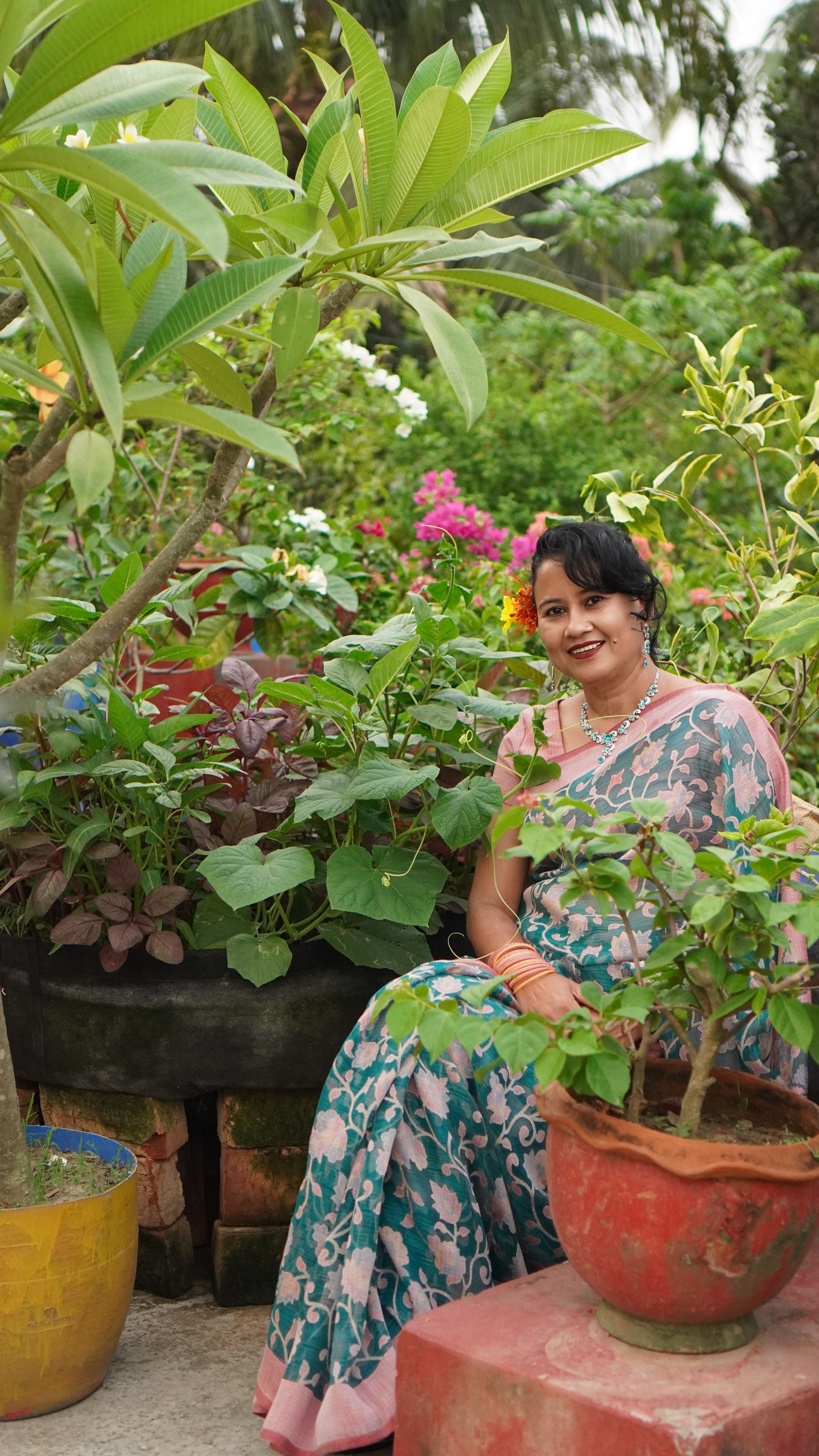 A smiling woman in a floral saree sits in a lush garden surrounded by vibrant plants.