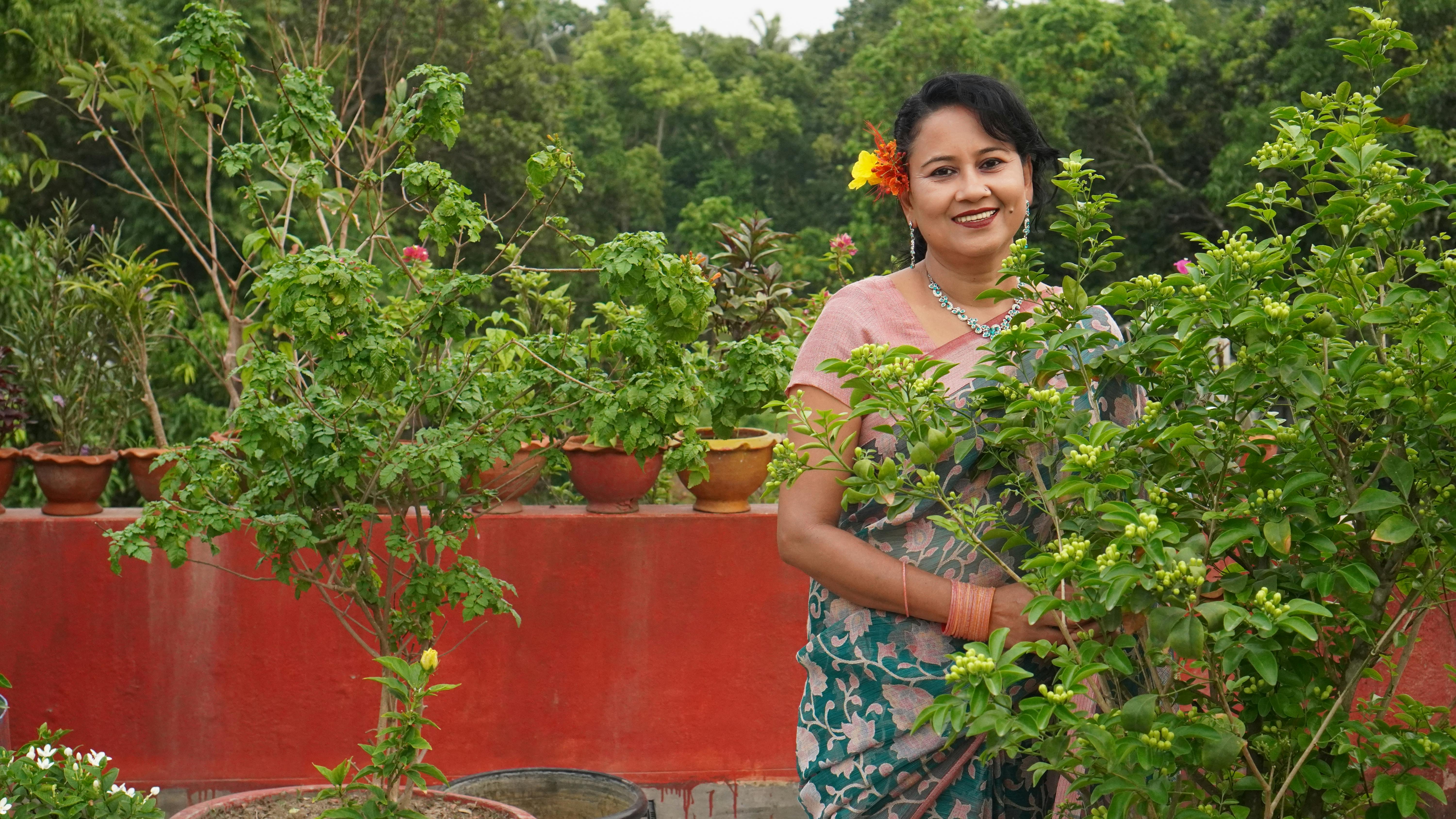 South Asian woman smiling among lush green plants in a vibrant garden setting.