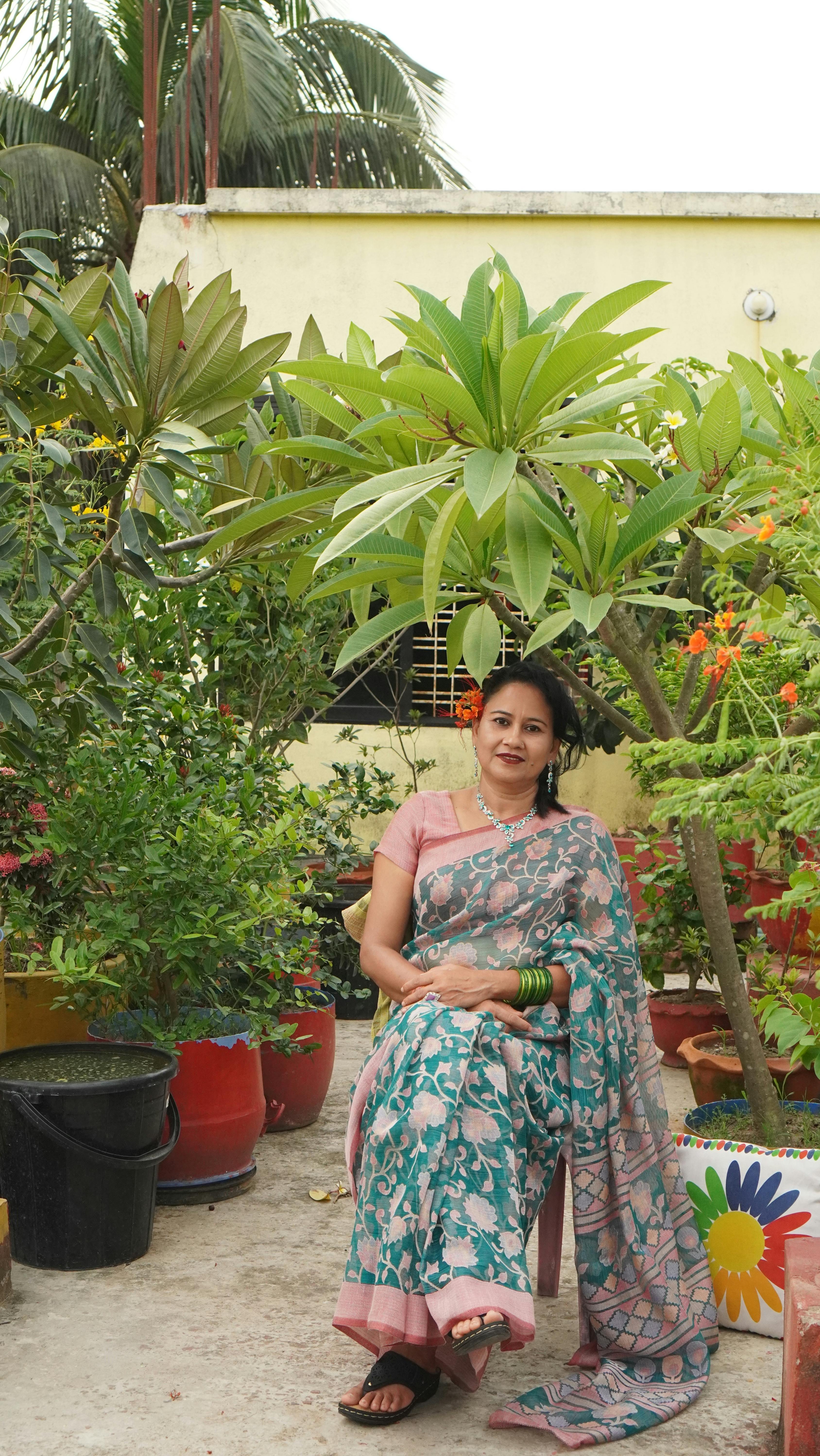 A woman in a traditional saree sitting in a garden surrounded by lush greenery.