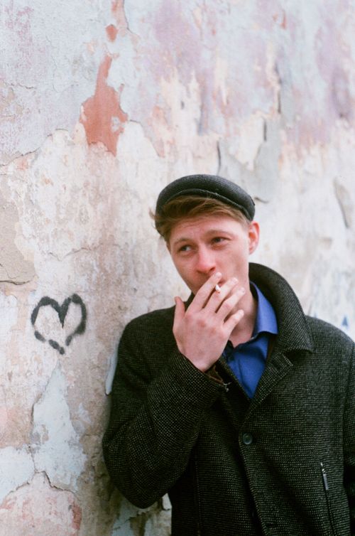 Free A young man wearing a flat cap smokes against a peeling wall with graffiti heart. Stock Photo