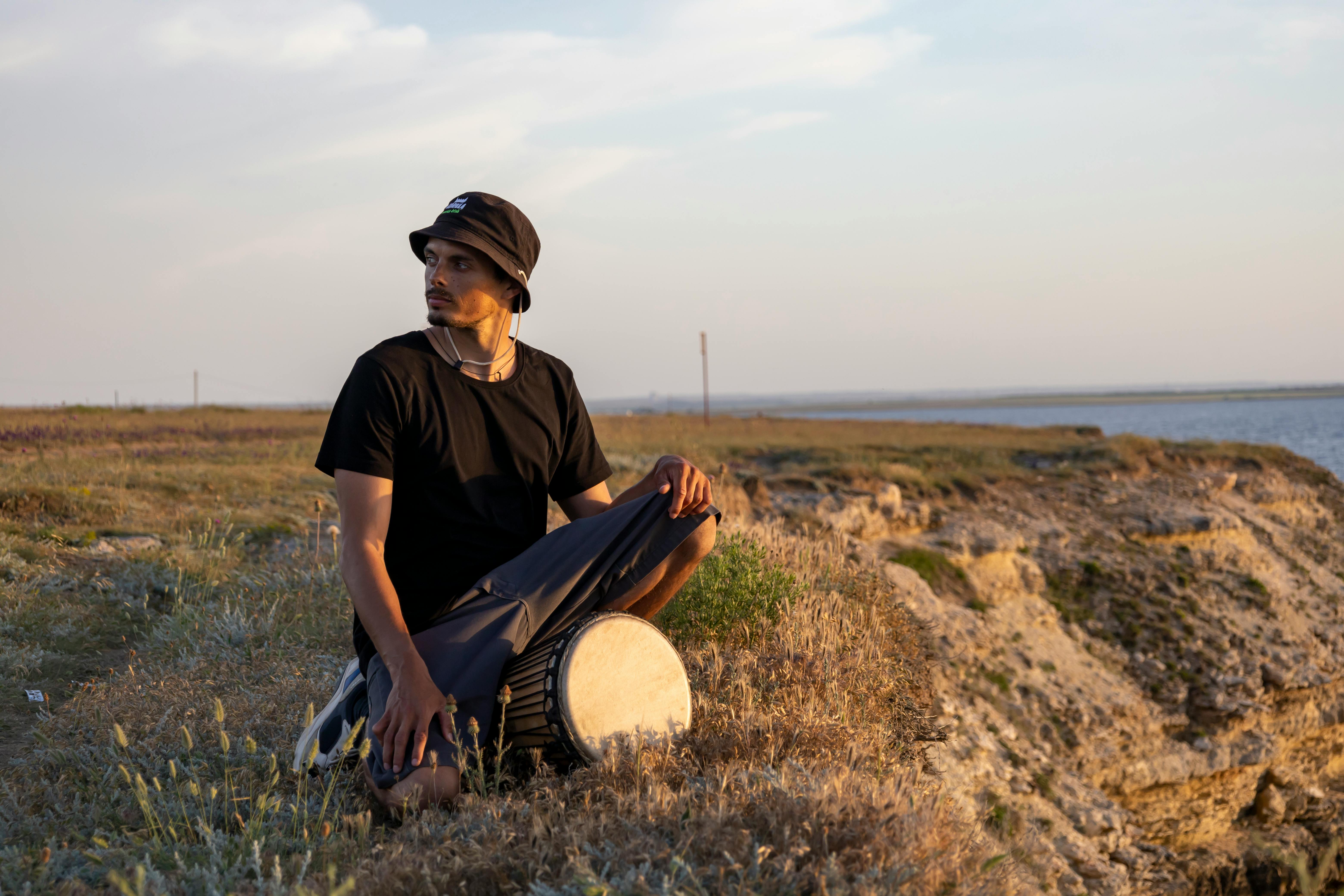 A man in a bucket hat sits with a drum on a grassy cliffside, gazing into the distance at sunset.