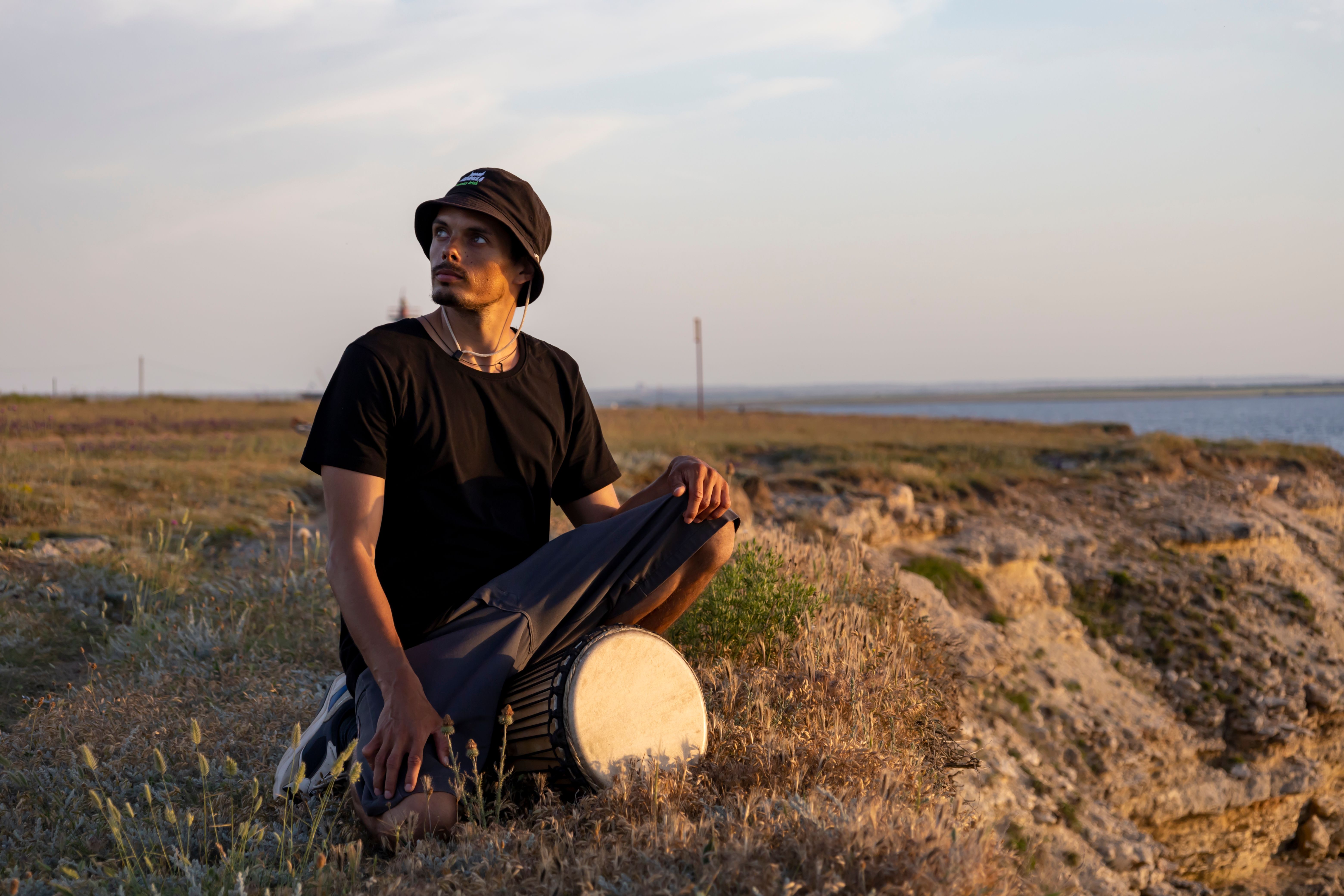 A young man with a drum sits on a cliff at sunset, gazing at the horizon.