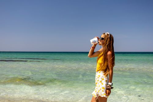 Free A woman in a yellow outfit drinks from a cup by the sea on a clear summer day. Stock Photo
