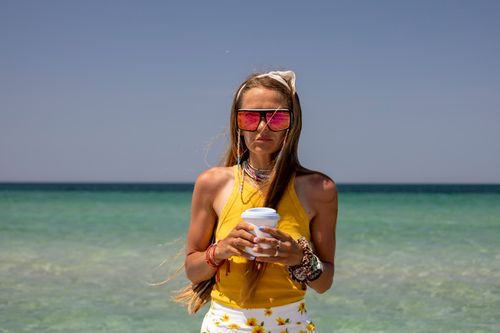 Free A woman in sunglasses holds a cup, enjoying a sunny day by the ocean. Stock Photo