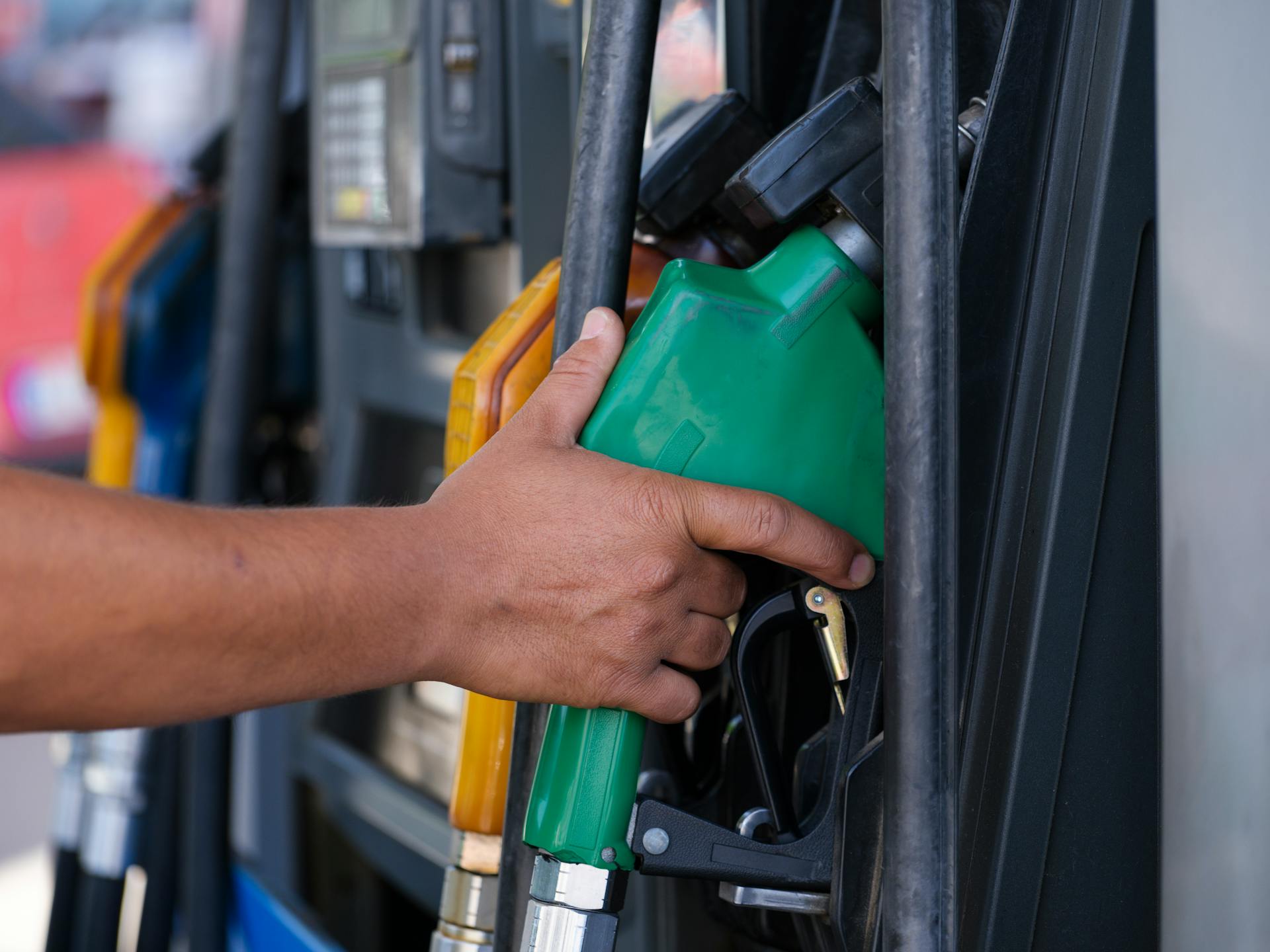 Close-up of a hand holding a green fuel nozzle at a gas pump station outdoors.