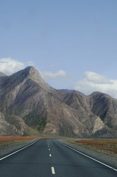 Scenic view of a straight highway leading to towering mountains under a clear blue sky and white clouds.