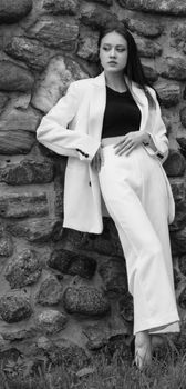 Fashionable woman in white suit leaning against a rustic stone wall in a monochrome setting.
