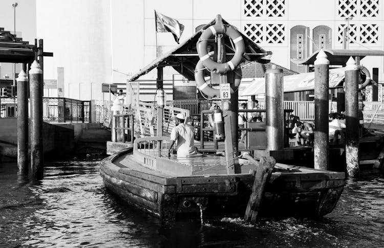 Black And White Photo Of A Man Sailing A Boat Towards A Harbour