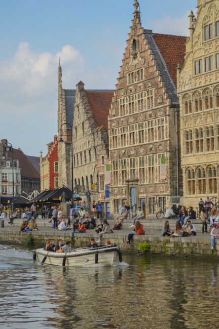 People enjoy a sunny day by the canal with historic European architecture and a passing boat.