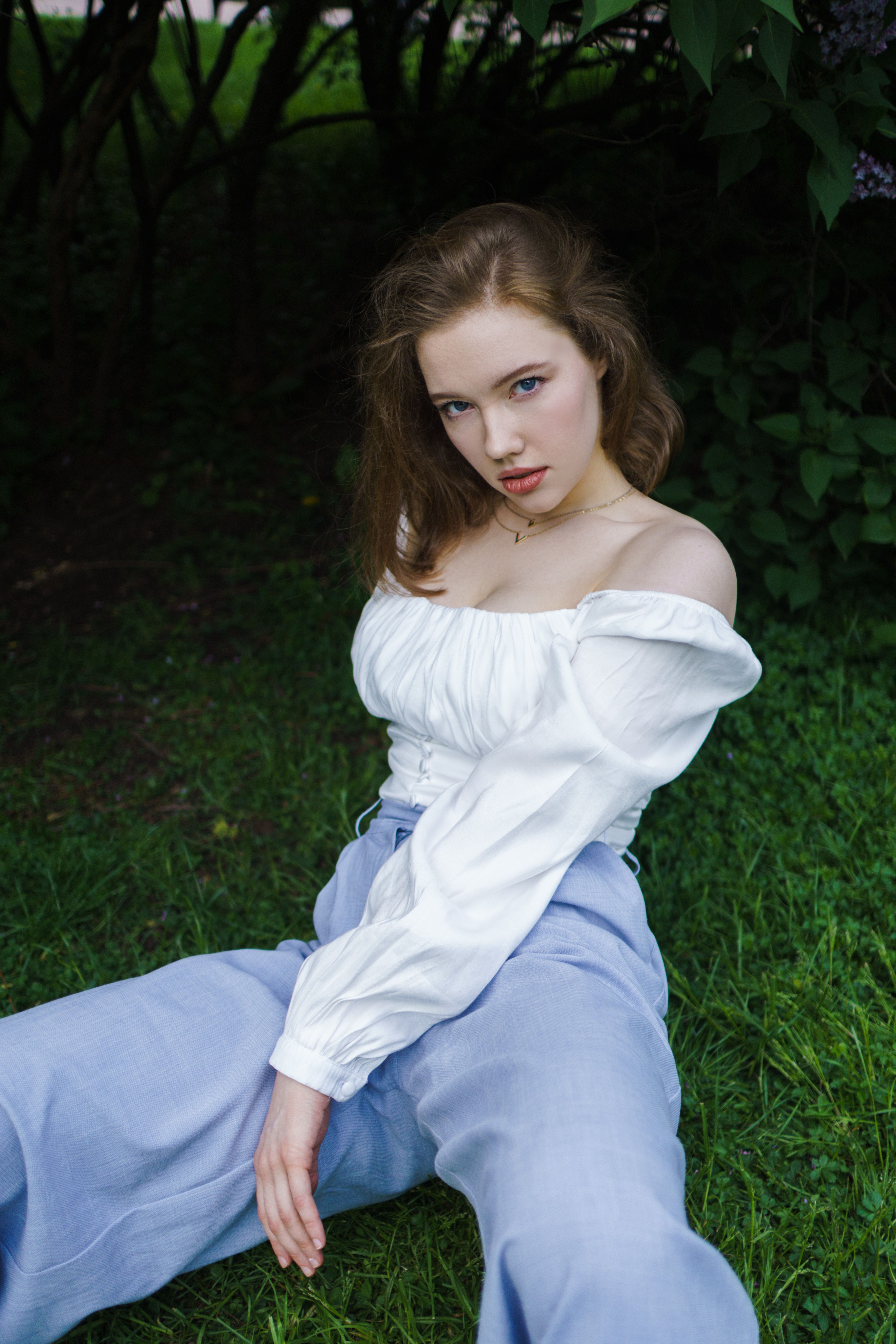 A young woman in white blouse and blue pants poses confidently on grass in a park setting.