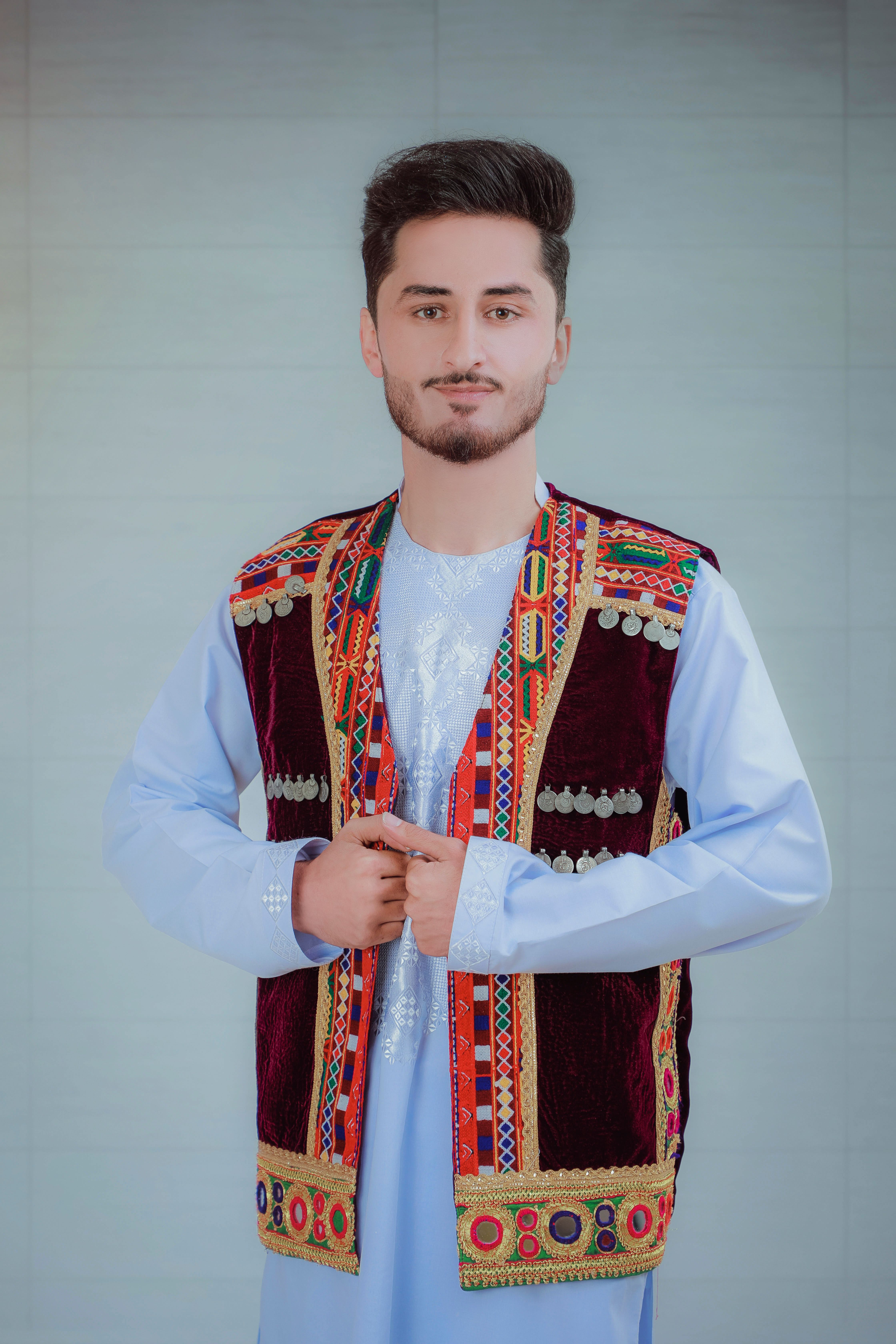 Young man wearing traditional embroidered vest and tunic, posing indoors with a neutral background.