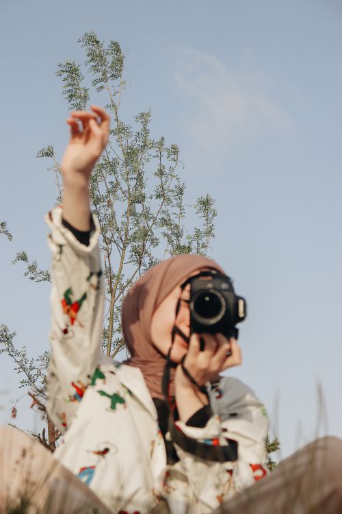 Free A Muslim woman with a camera takes photos in a natural setting with a blue sky backdrop. Stock Photo