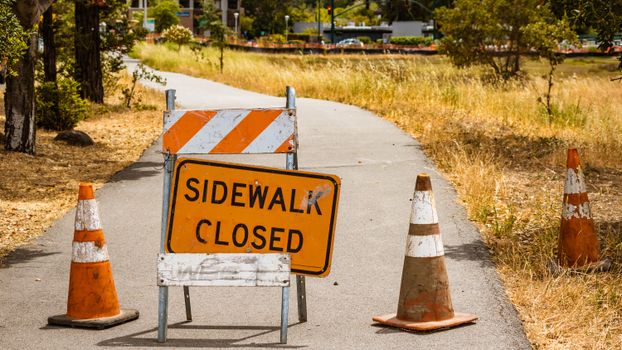 Photo of Sidewalk a Closed Sign Askew on a Stand and Three Road Cones against the Background of Trees and Grass