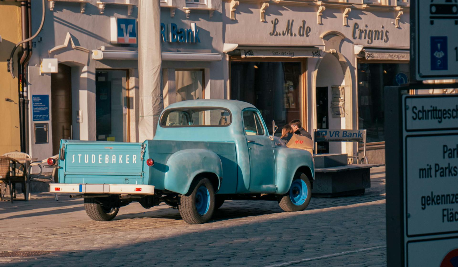 A classic Studebaker pickup truck is parked on a sunlit cobblestone street outside a bank in Europe.