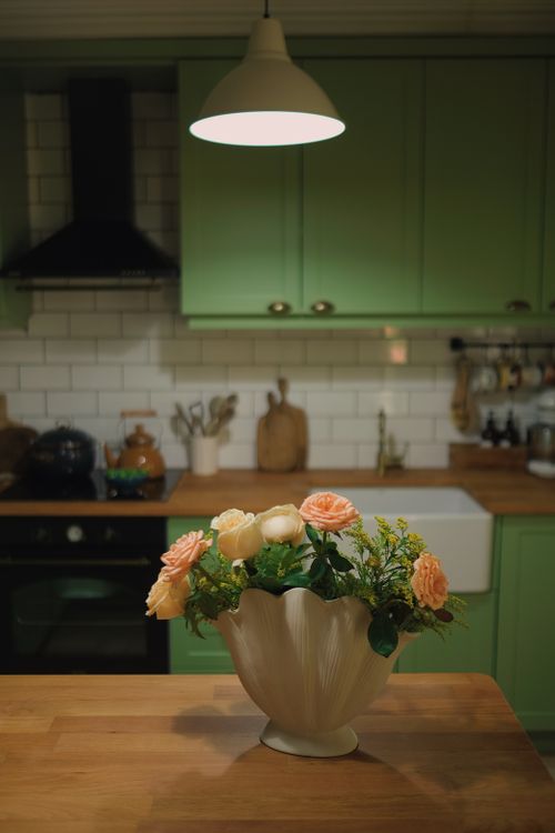 Free A cozy kitchen interior with a floral arrangement under warm lighting, featuring pastel green cabinets. Stock Photo