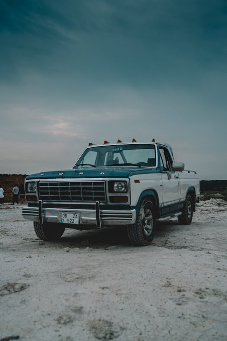 Blue And White Ford Pickup Truck On A Beach Under A Cloudy Sky