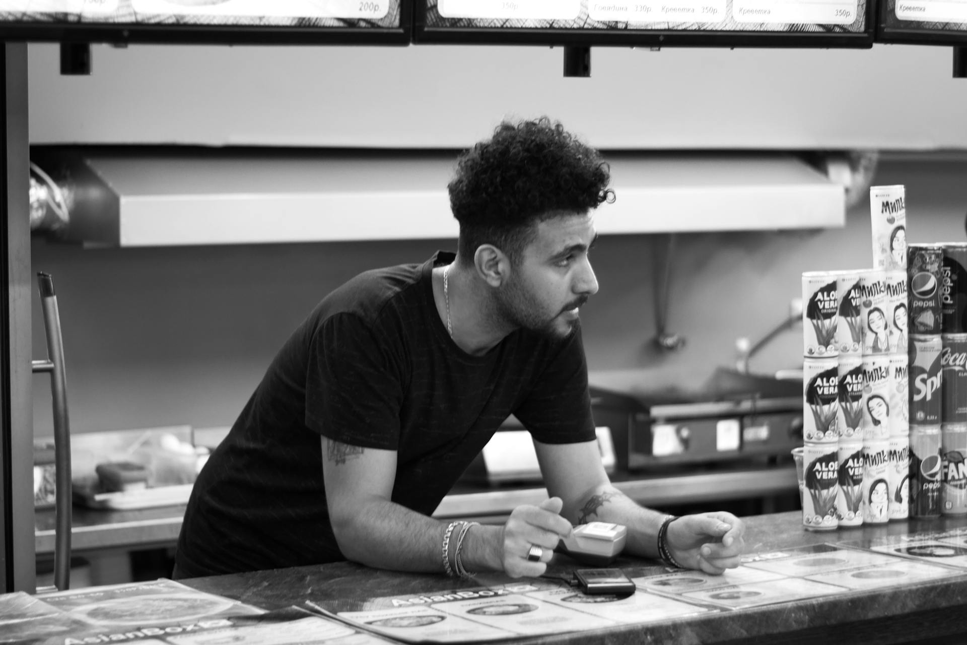 Black and white photo of a man in a shop setting, leaning on the counter with beverages displayed.