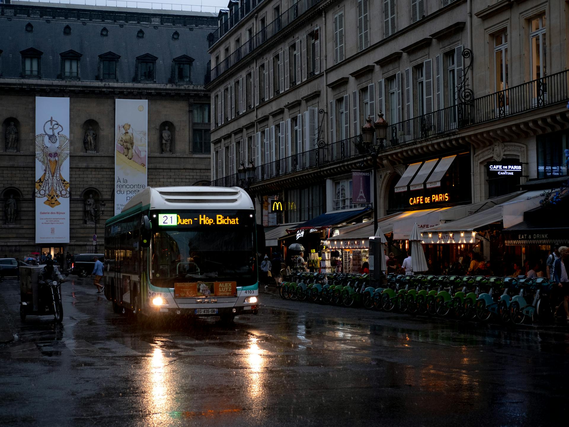 A bus navigates wet Paris streets at dusk, with nearby cafes and architecture adding charm.