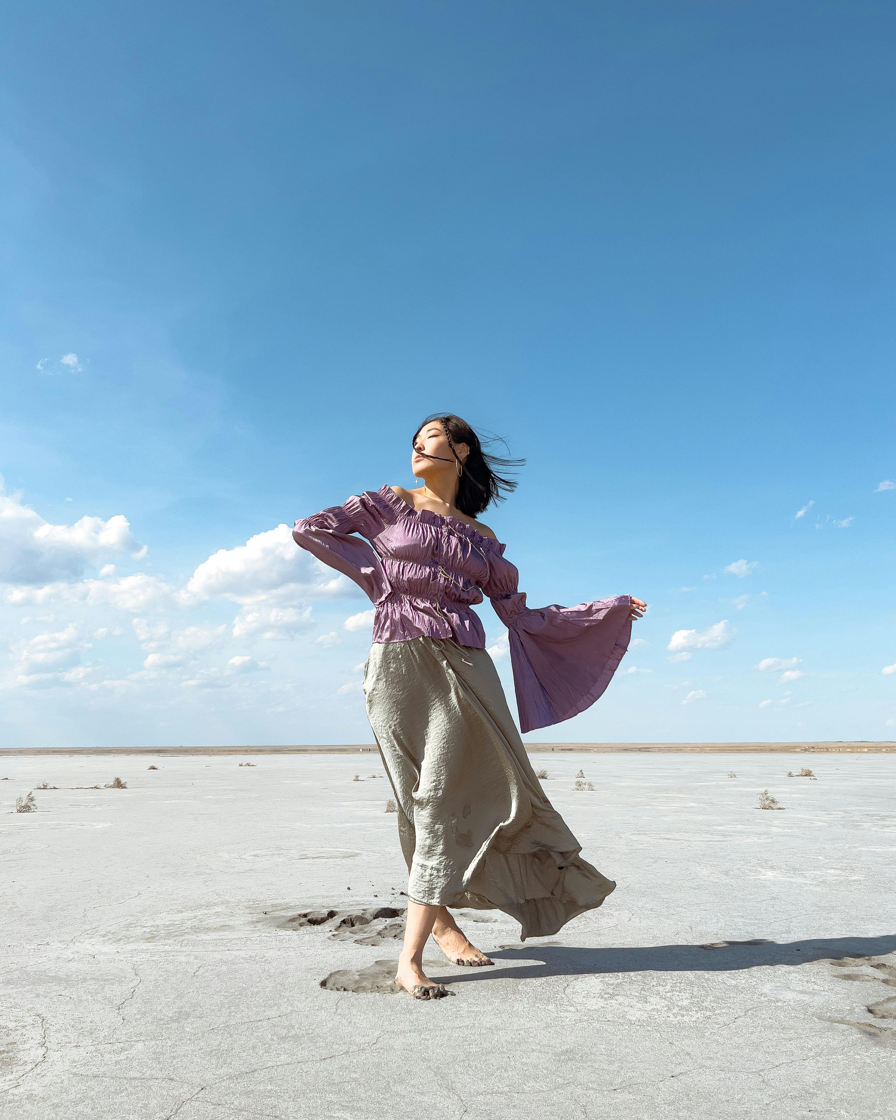 A woman in flowing garments dancing gracefully in a sunny desert landscape under a blue sky.