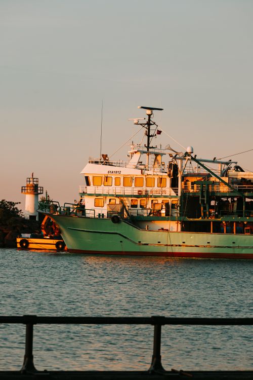 Free A large ship docked at a harbor during a warm, glowing sunset with a lighthouse in the background. Stock Photo