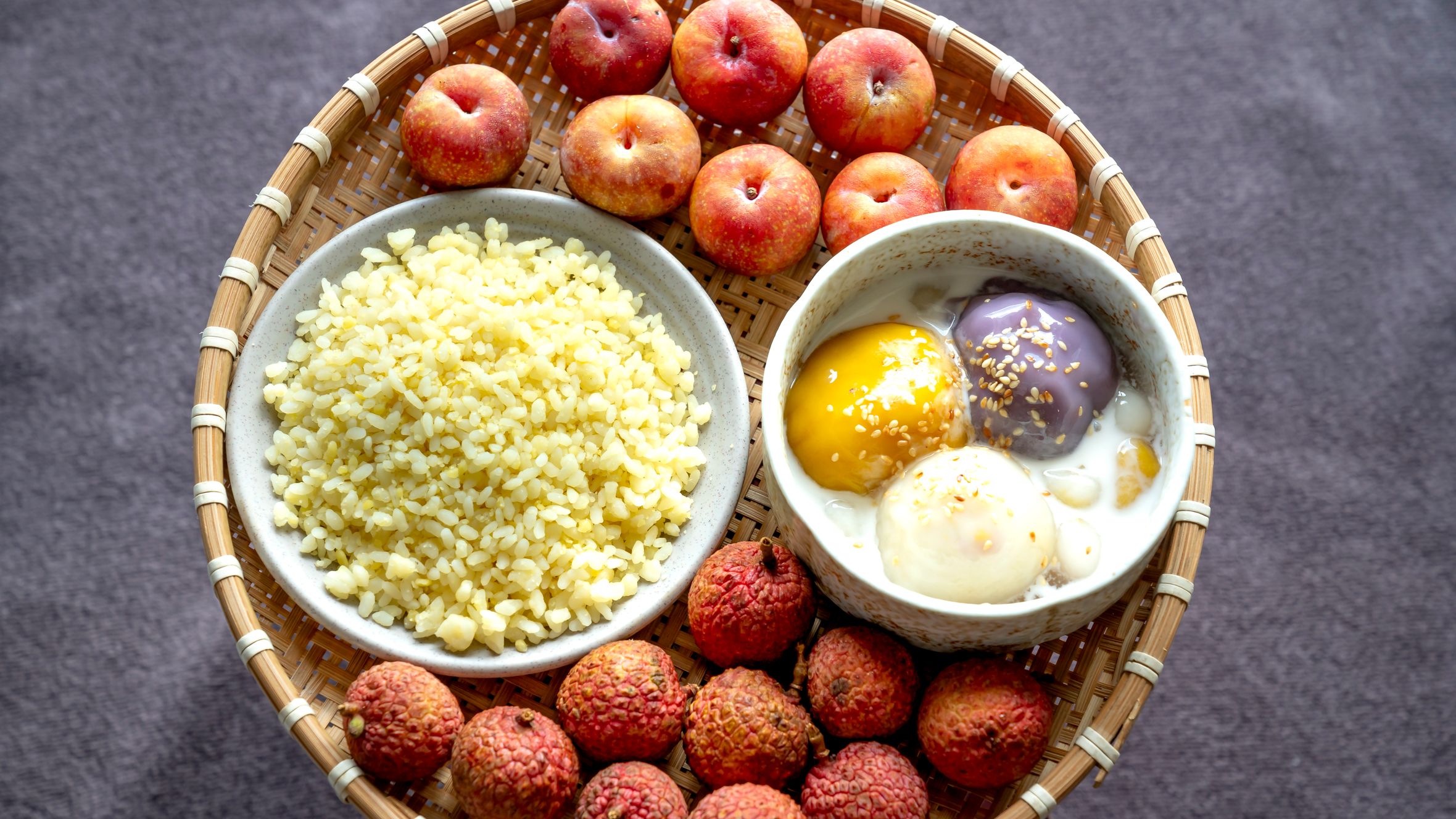 A vibrant top view of a woven basket with lychees, plums, rice, and desserts.