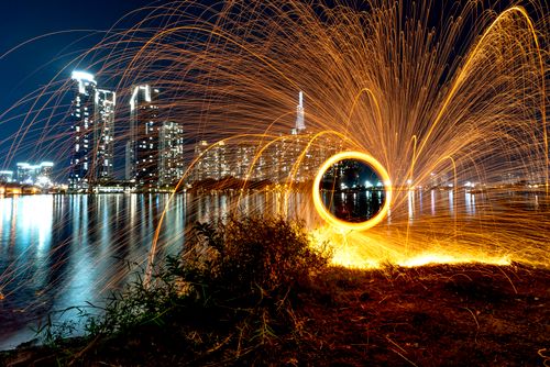 Free Captivating steel wool photography creates vibrant sparks near a glowing city skyline reflected in tranquil water. Stock Photo