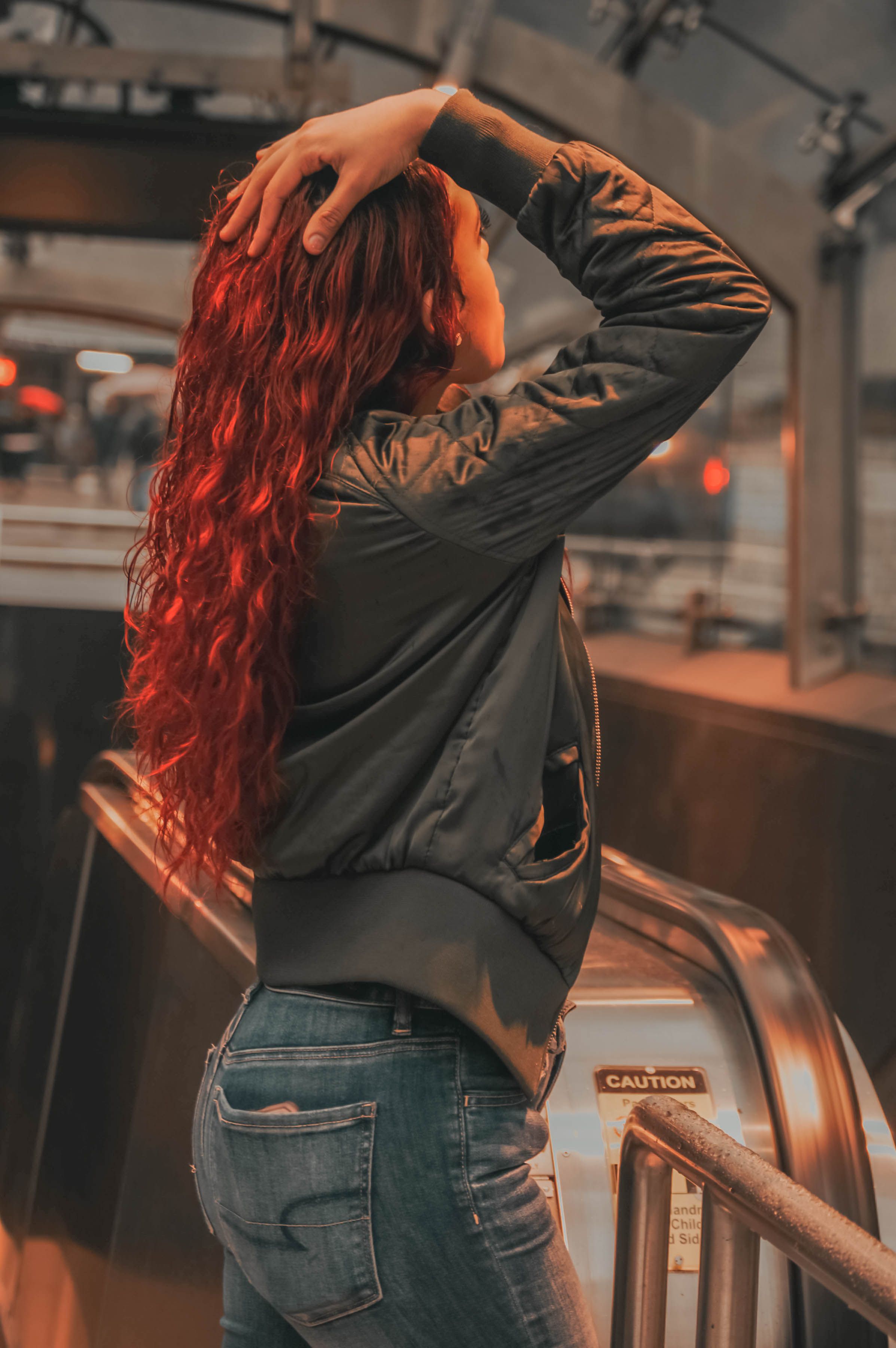 A woman with long red hair poses indoors near an escalator, touching her head.