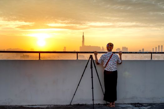 A woman stands with a camera tripod on a rooftop, viewing a city skyline at sunset.