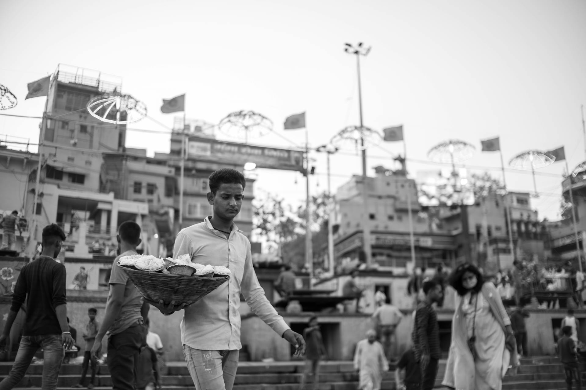 Monochrome photo of a street vendor carrying goods in a busy Indian market with crowd.