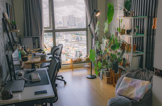 Cozy home office in urban apartment with lush plants and city view through large window.
