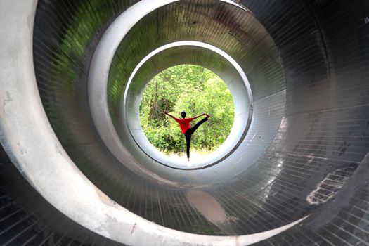An individual in a vibrant pose within a large outdoor circular pipe, surrounded by nature.