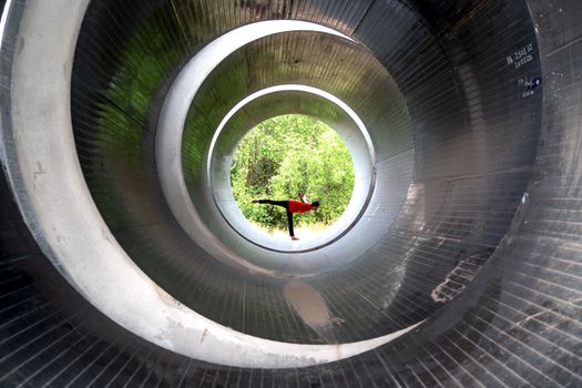 A person is performing a yoga pose inside a large circular metal tunnel with trees visible in the background.