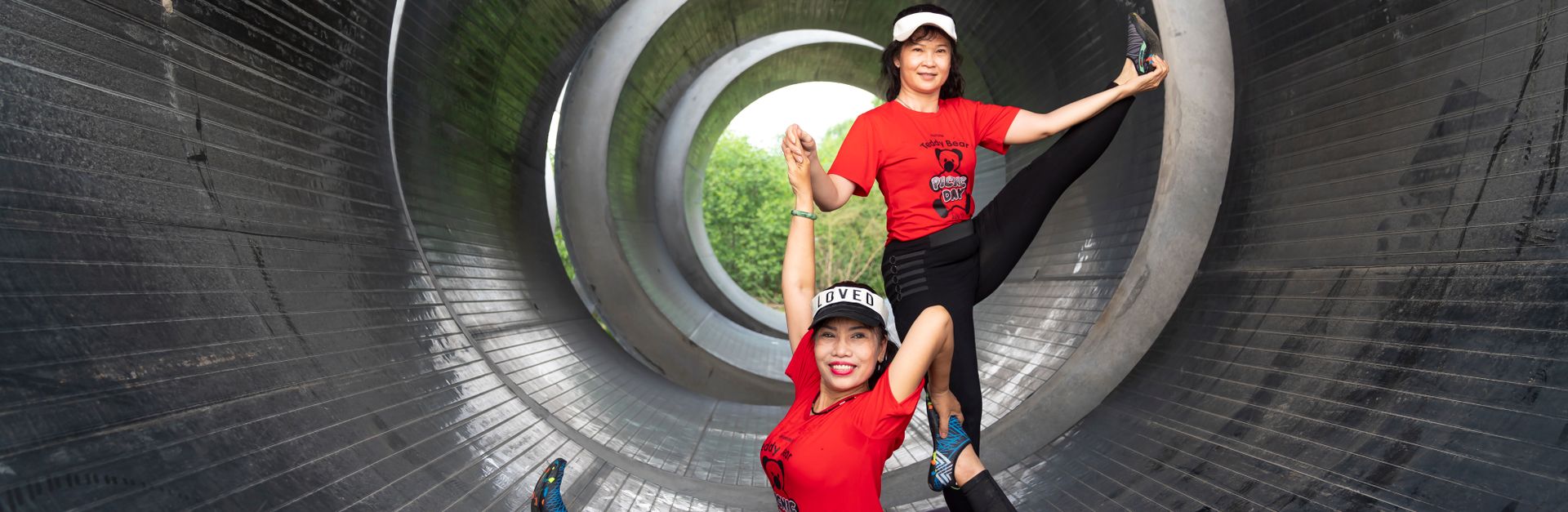 Two women perform stretching exercises inside a large metal tunnel, displaying flexibility.