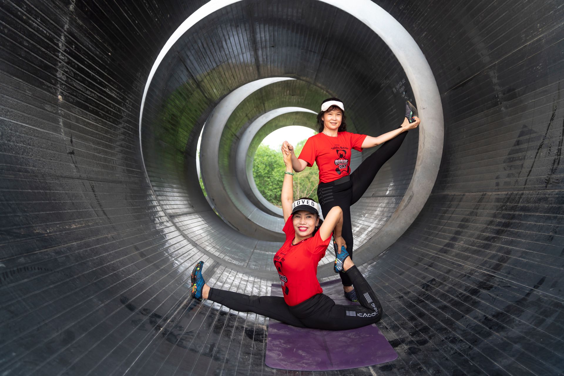 Two women perform stretching exercises inside a large metal tunnel, displaying flexibility.