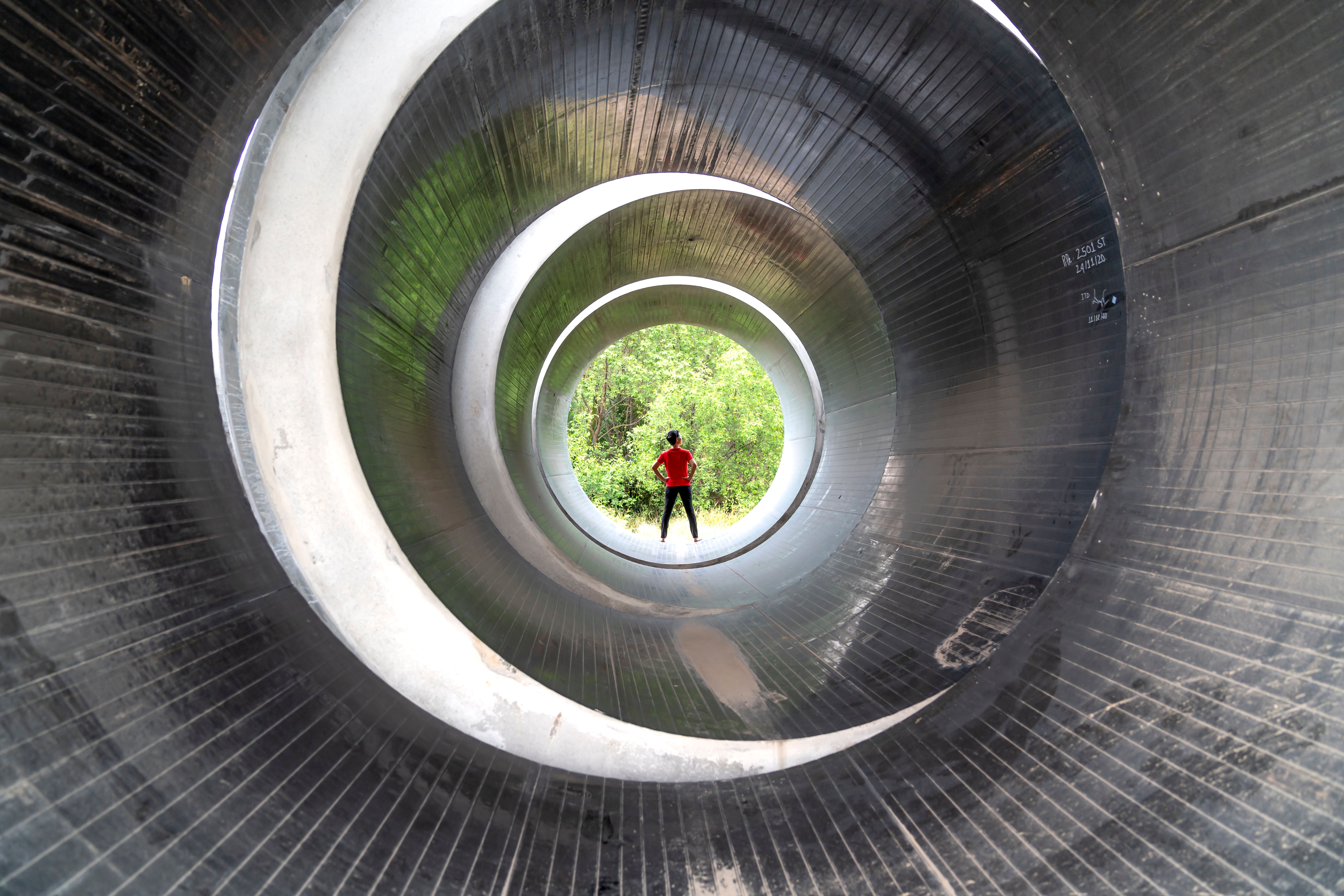 Silhouette of a person in a red shirt inside a massive industrial pipe with greenery in the distance.