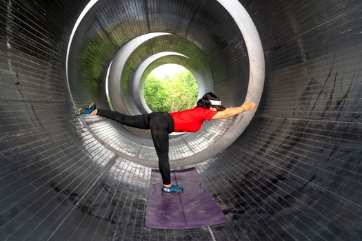 Female athlete practices balance and stretching inside an industrial concrete pipe using a yoga mat.
