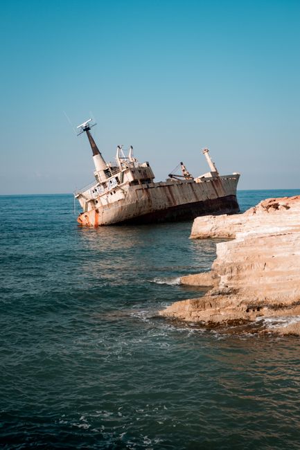 A rusty shipwreck remains lodged on the rocky coastline, surrounded by calm sea waters under a clear sky.