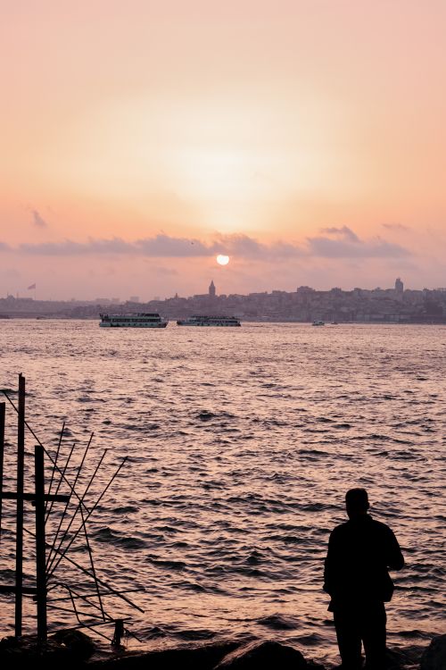 Free A man silhouetted against a vibrant sunset by the sea, with distant cityscape view. Stock Photo