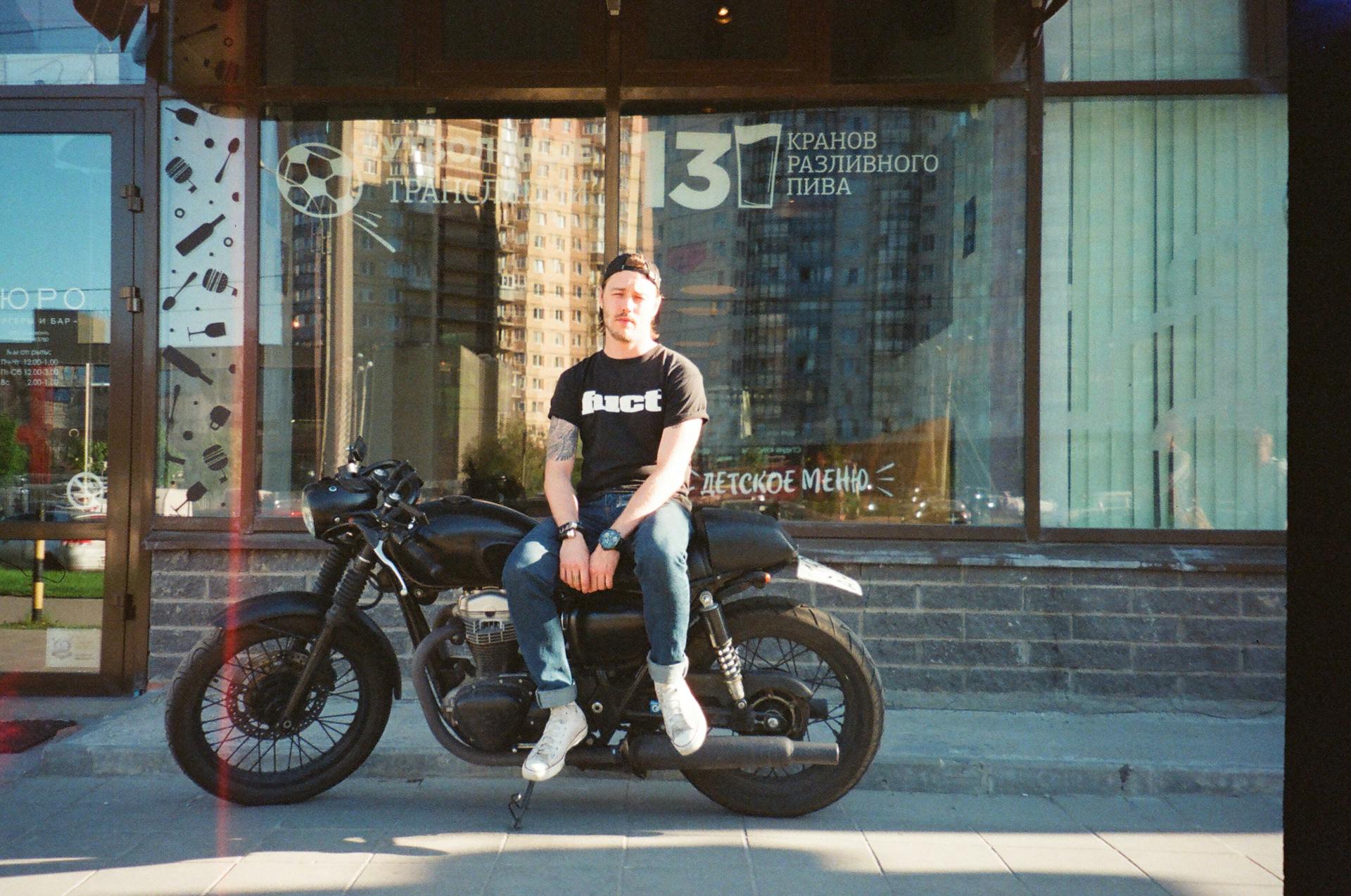 A stylish young man sits on a vintage motorcycle in front of a modern building, showcasing urban lifestyle.