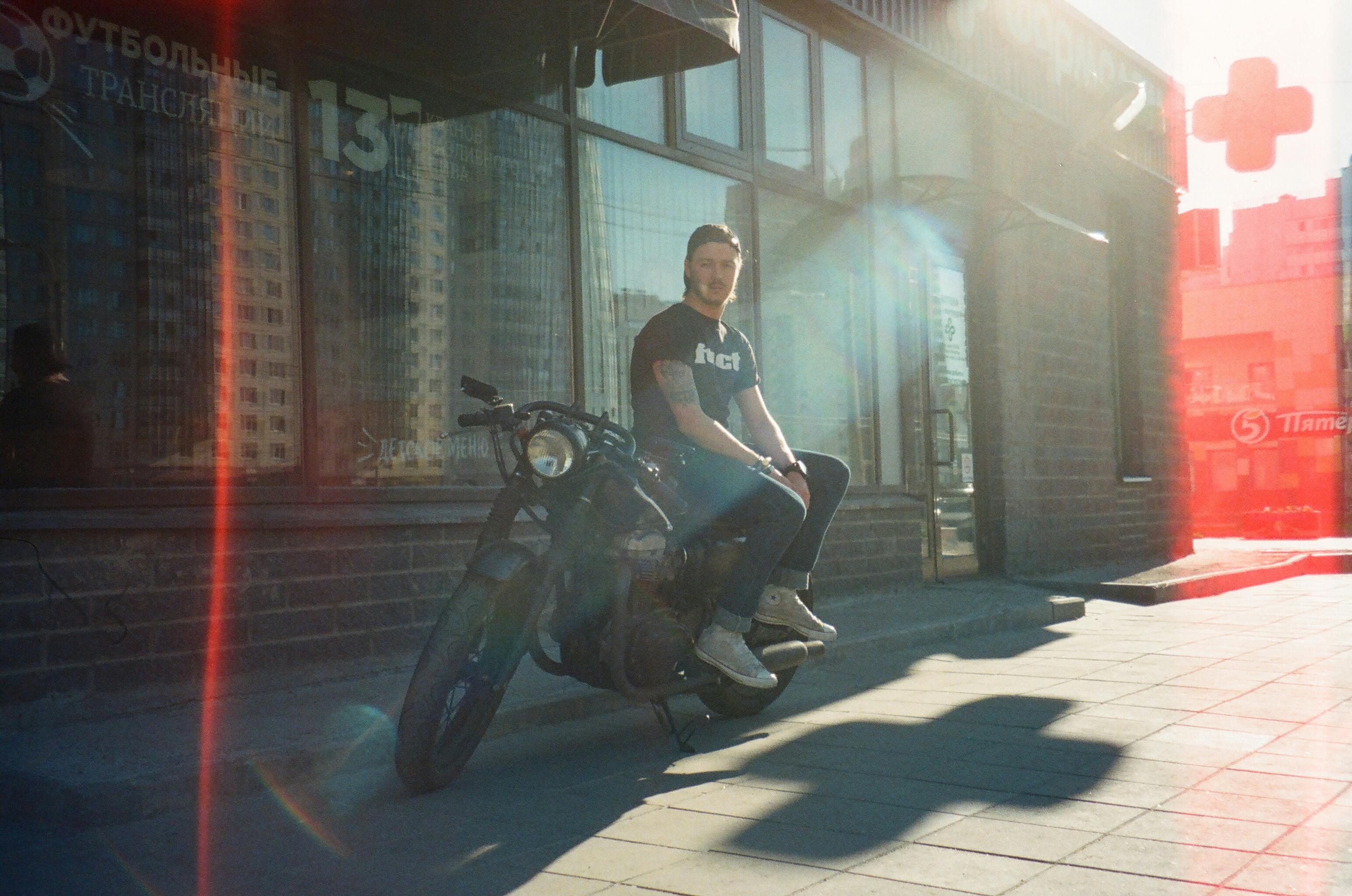 Man sitting on a motorcycle in urban street during daylight, showcasing casual fashion and urban lifestyle.